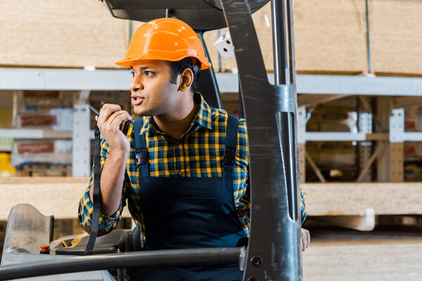 indian warehouse worker in helmet talking on walkie talkie while sitting in forklift machine
