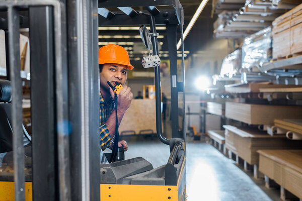 handsome indian worker sitting in forklift machine in warehouse and talking on walkie talkie