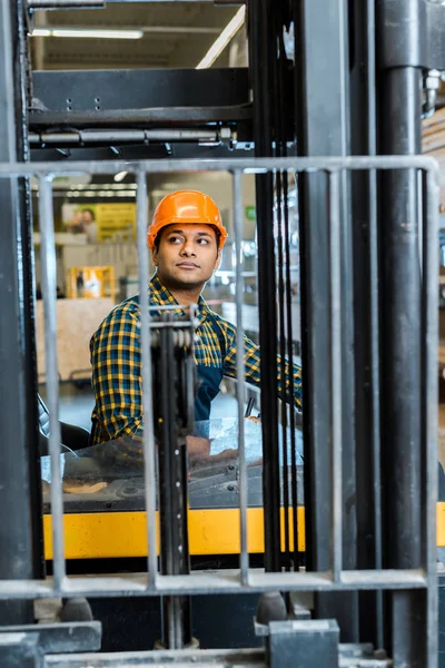 handsome, attentive indian worker sitting in forklift machine in warehouse