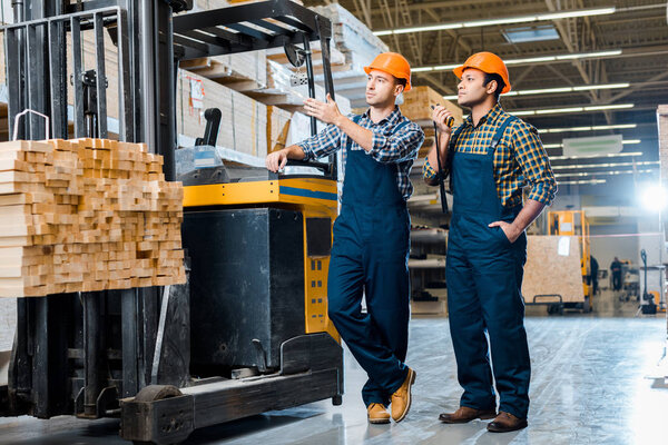 handsome multicultural warehouse workers in helmets standing near forklift machine in warehouse