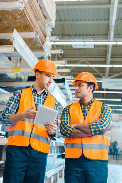 multicultural warehouse workers in safety vasts and helmets talking while using digital tablet
