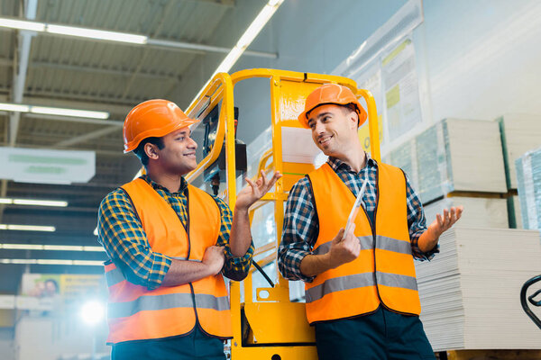 cheerful multicultural workers talking and gesturing while standing near scissor lift 