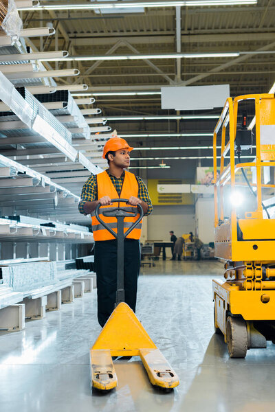 handsome indian worker in uniform standing near pallet jack in warehouse