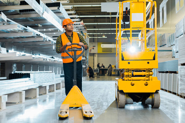 serious indian worker in safety vest and helmet standing near pallet jack in warehouse