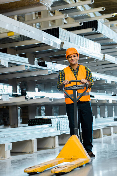 cheerful indian worker standing near pallet jack, smiling and looking at camera