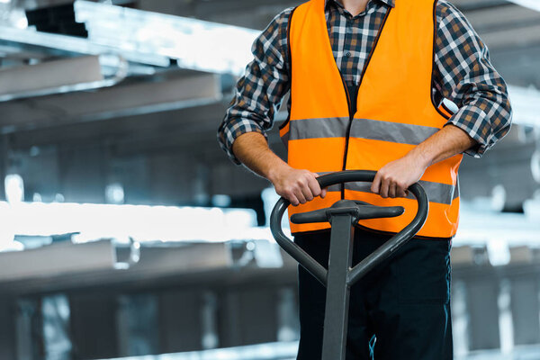 partial view of warehouse worker in safety vast standing with pallet jack