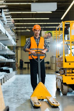 cheerful warehouse worker standing with pallet jack, smiling and looking at camera clipart