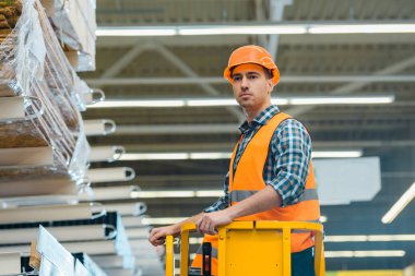 handsome, serious worker standing on scissor lift in warehouse clipart