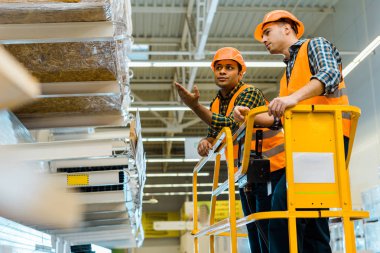 serious indian worker pointing with hand at shelf with construction materials while standing on scissor lift with colleague clipart