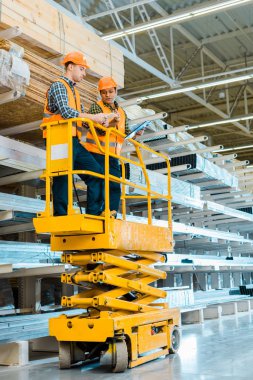 multicultural warehouse workers with digital tablet and clipboard standing on scissor lift  clipart