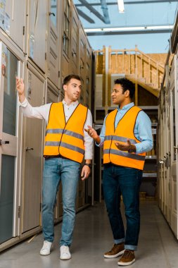 smiling multicultural coworkers in safety vests talking in in doors department clipart