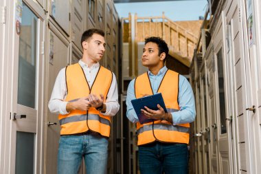 serious multicultural warehouse workers talking while standing in doors department clipart