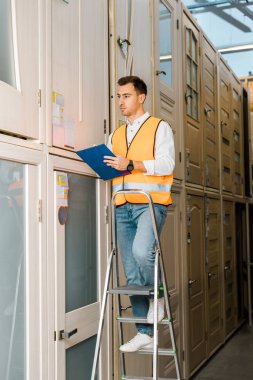 handsome, serious warehouse worker with clipboard standing on ladder in doors department clipart