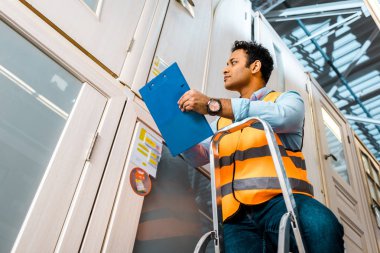 handsome, concentrated indian warehouse worker standing on ladder and holding clipboard clipart
