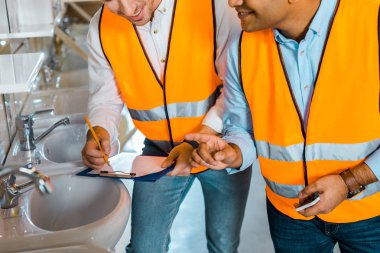 cropped view of multicultural colleagues in safety vests working in plumbing department clipart