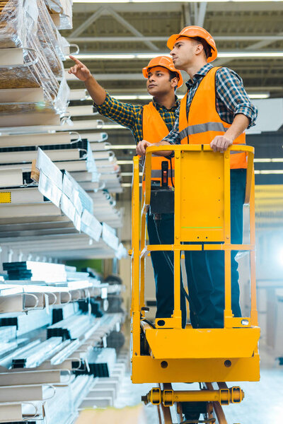 serious indian worker pointing with finger at construction materials while standing on scissor lift near colleague 
