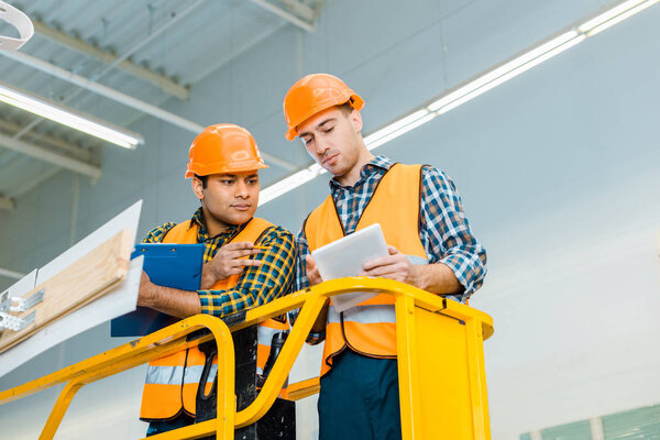 thoughtful multicultural workers with digital tablet and clipboard standing on scissor lift