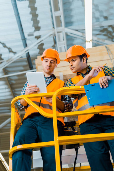 serious multicultural warehouse workers using digital tablet while standing on scissor lift 