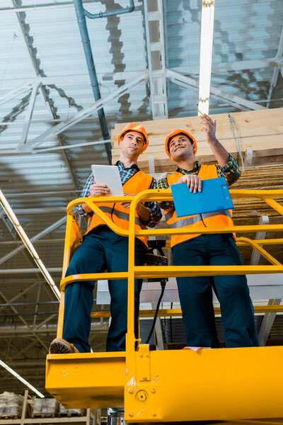 smiling multicultural workers with digital tablet and clipboard standing on scissor lift in warehouse 