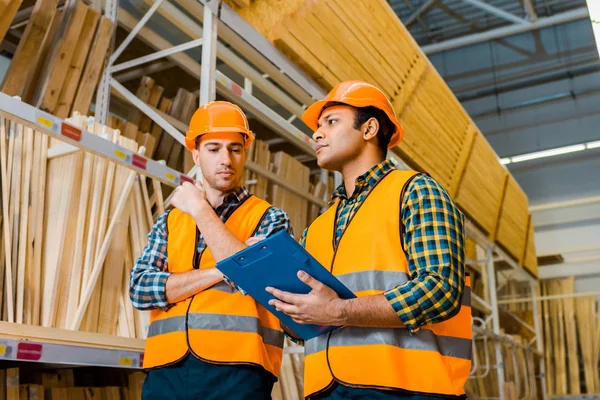 concentrated multicultural workers with clipboard standing near racks with wooden construction materials