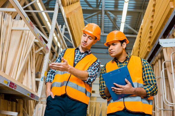 handsome multicultural worker pointing with hand at wooden construction materials near indian colleague