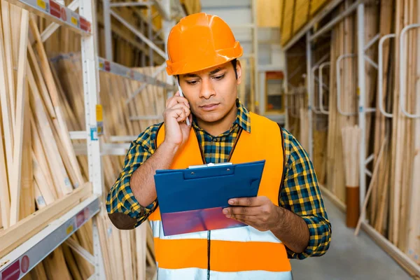 concentrated indian worker talking on smartphone and looking at clipboard