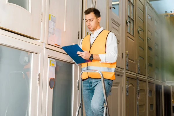 selective focus of concentrated worker with clipboard standing on ladder in warehouse