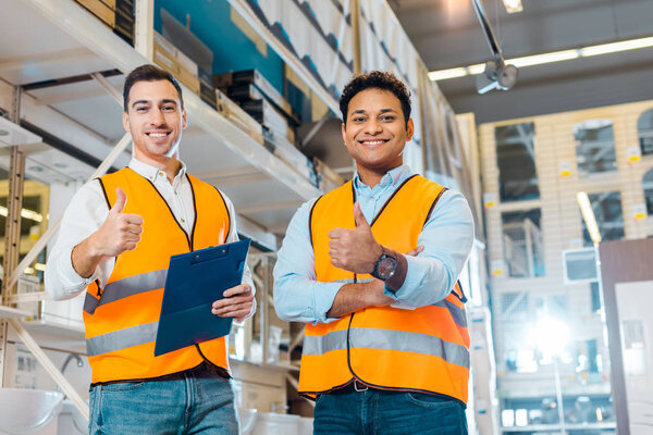 cheerful multicultural warehouse workers showing thumbs up and looking at camera