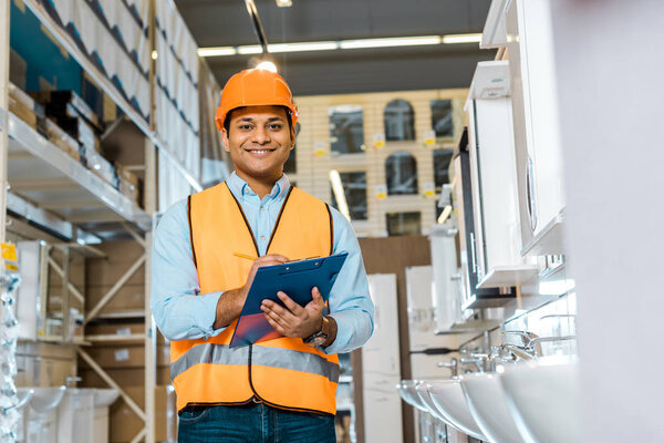 cheerful indian warehouse worker writing in clipboard, smiling and looking at camera  