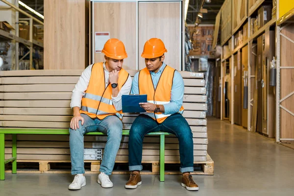 Concentrated Multicultural Workers Sitting Bench Warehouse Looking Clipboard — Stock Photo, Image