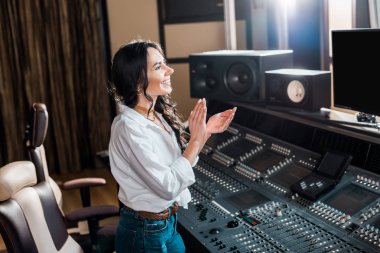 attractive, smiling sound producer applauding in recording studio near mixing console  clipart