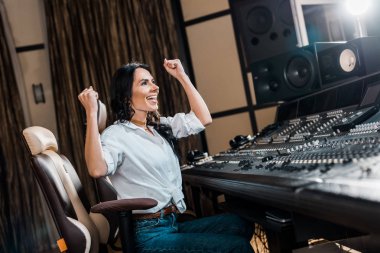 cheerful sound producer showing winner gesture sitting near mixing console in recording studio clipart
