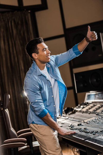 smiling mixed race sound producer showing thumb up while working at mixing console