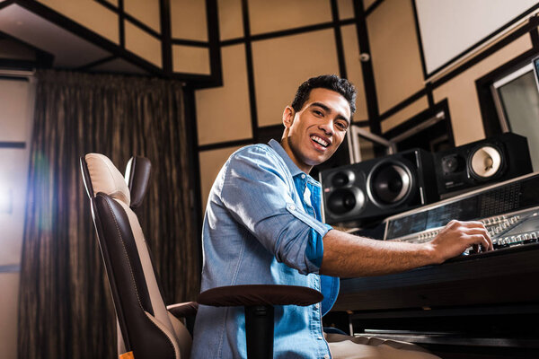 cheerful mixed race sound producer sitting in office chair in recording studio and looking at camera