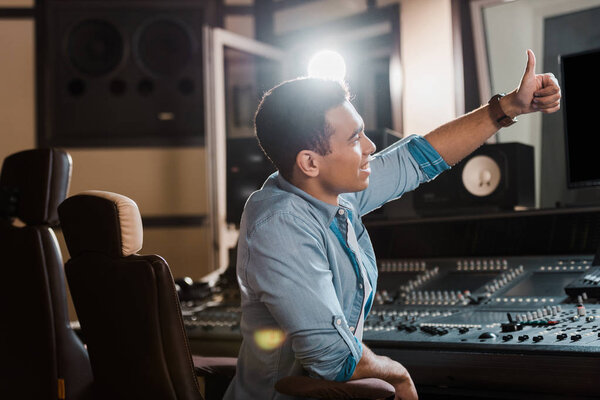 smiling mixed race sound producer showing thumb up while working in recording studio