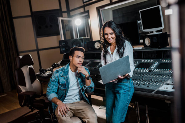 smiling pretty sound producer using laptop near mixed race friend in recording studio