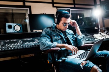concentrated mixed race sound producer in headphones using laptop in recording studio clipart