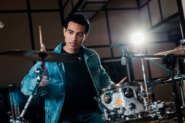 handsome mixed race musician playing drums in dark recording studio clipart
