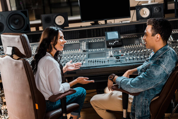 two smiling multicultural sound producers sitting by mixing console in recording studio