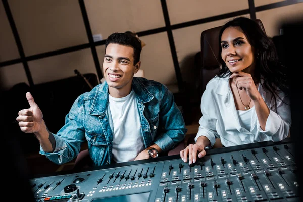 handsome multicultural sound producer showing thumb up while sitting near smiling attractive colleague