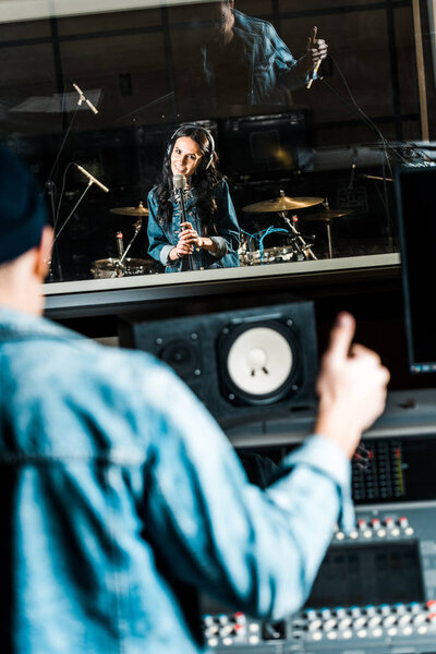 selective focus of mixed race sound producer showing thumb up to beautiful singing girl