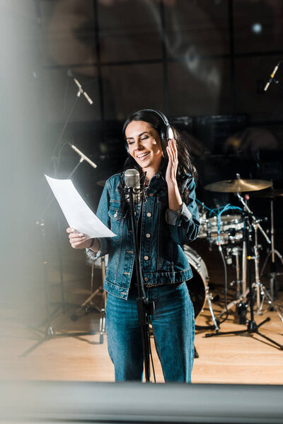 selective focus of cheerful woman in headphones singing in recording studio