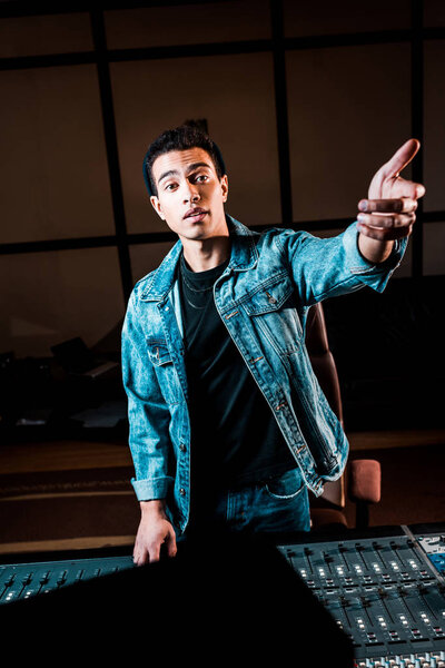 handsome mixed race sound producer gesturing while standing near mixing console in recording studio