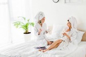 elegáns nők fürdőköpenyek, fülbevalók és törülközők a fejek gazdaság kávéscsészét és csészealjak, miközben beszél az ágyban