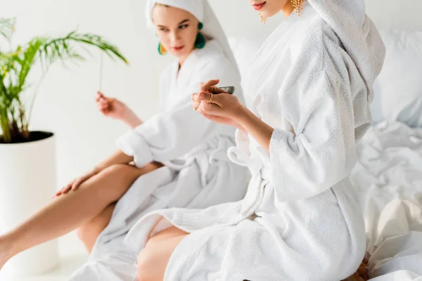 selective focus of stylish women in bathrobes and jewelry, with towels on heads sitting on bed and filing nails near green plant