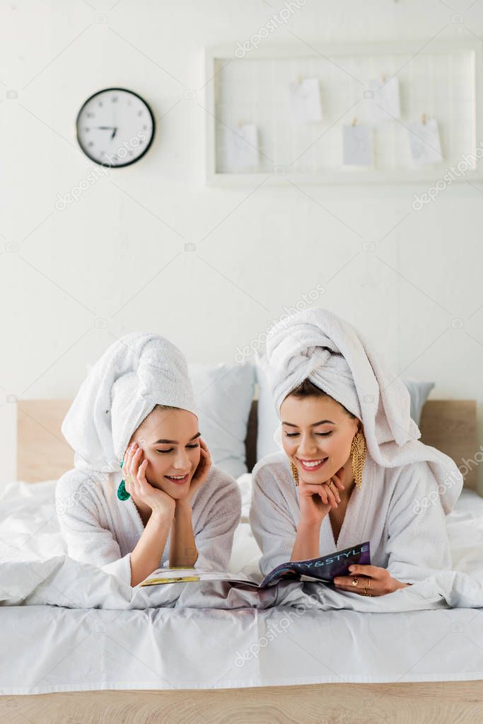 happy stylish women in bathrobes, earrings and with towels on heads reading magazine while lying in bed