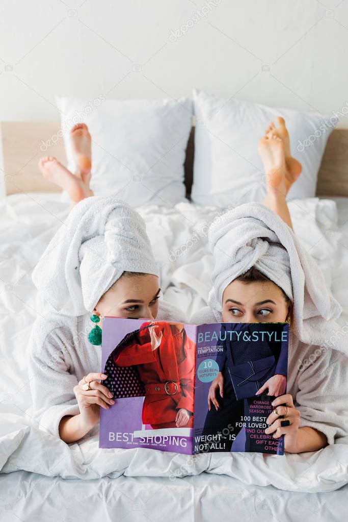 overhead view of stylish barefoot women in bathrobes, earrings and with towels on heads hiding behind magazine together while lying in bed