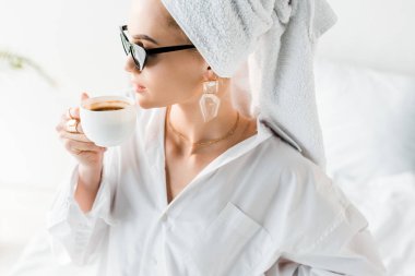 Gömlekli, güneş gözlüklü, mücevherli ve kafasında havluyla kahve içen şık bir kadın.