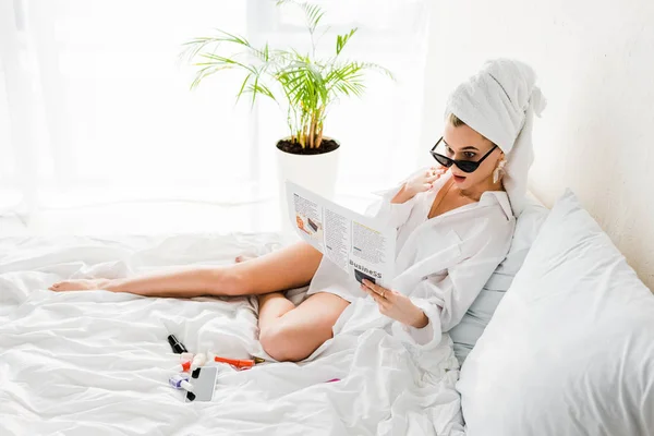 シャツ サングラス ジュエリーを着たスタイリッシュなショックを受けた女性が マニキュアやスマートフォンの近くに新聞を持ってベッドに横たわっている — ストック写真