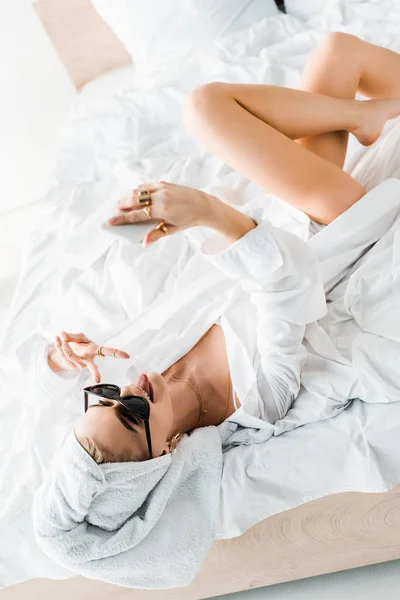 シャツ サングラス ジュエリー タオルを頭に付けて舌を出しながらスマートフォンでセルフィーを撮り ピースサインを見せ ベッドに横たわっているスタイリッシュな女性 — ストック写真